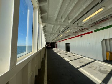 Feribotun içi geniş bir koridor, geniş pencereler, parlak tepe ışıkları, ve zeminde yansımalar, sakin bir atmosfer yaratıyor.