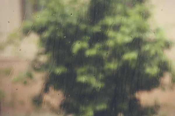 View to raining weather through the home window. Raindrops on the window glass. Heavy raining outdoors. Spring rain season