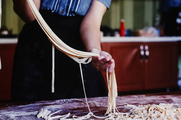 Νεαρή νοικοκυρά ετοιμάζει τα σπιτικά ζυμαρικά στην κουζίνα. Γυναικεία χέρια που κρατούν μακριά λωρίδα ζύμης για το μαγείρεμα ζυμαρικών. — Φωτογραφία Αρχείου