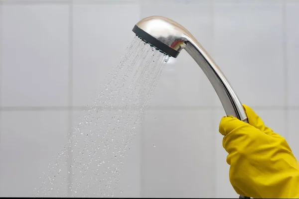 A person in rubber gloves holding showerhead with flowing water. Women in yellow protective gloves holding the head of the shower with running water in the bath — Stock Photo, Image