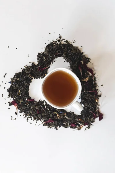 Kształt serca wykonane z mieszaniną różnych suszonych liści herbaty i filiżankę czarnej herbaty — Zdjęcie stockowe