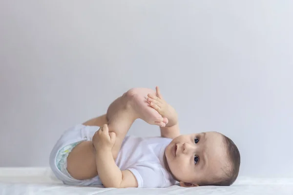 Petit garçon affamé qui prend les pieds dans la bouche. Bébé de 6 mois s'amusant dans la literie blanche. Bébé mignon couché sur le lit. Famille, nouvelle vie, concept d'enfance . — Photo