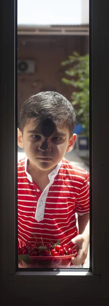 Un niño pequeño parado afuera con unas cerezas y mirando por la puerta. Infancia, concepto de verano — Foto de Stock
