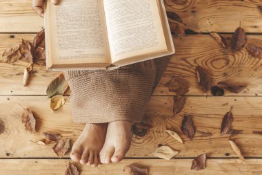 Reading book. Female sit at wooden floor with fall leaves. Woman covered with knitted plaid and reading interesting book clipart
