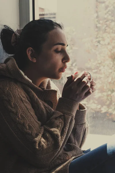 Junge brünette Frau trinkt eine Tasse Tee. Frau, die zu Hause am Fenster sitzt. Kalte Herbsttage. Fallrückzieher. — Stockfoto