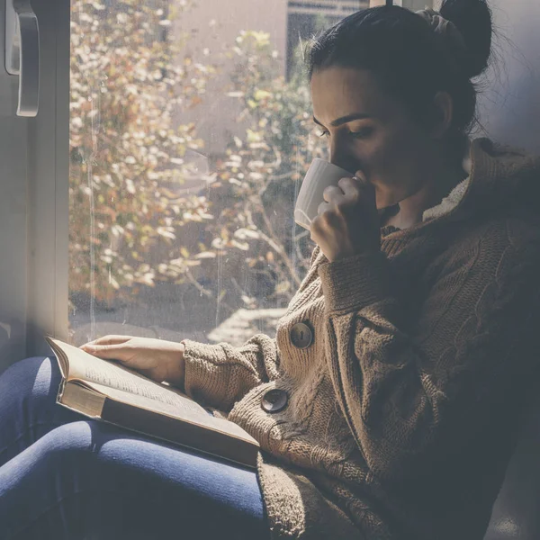 Jonge brunette vrouw drinken een kopje thee of koffie en het lezen van een boek. Vrouw zittend thuis bij het raam en lees een boek — Stockfoto