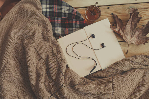Осенний фон. Открытая книга, осенний лист и черные наушники на вязаном свитере. Вязаный шарф на деревянном столе. Концепция осенних выходных

