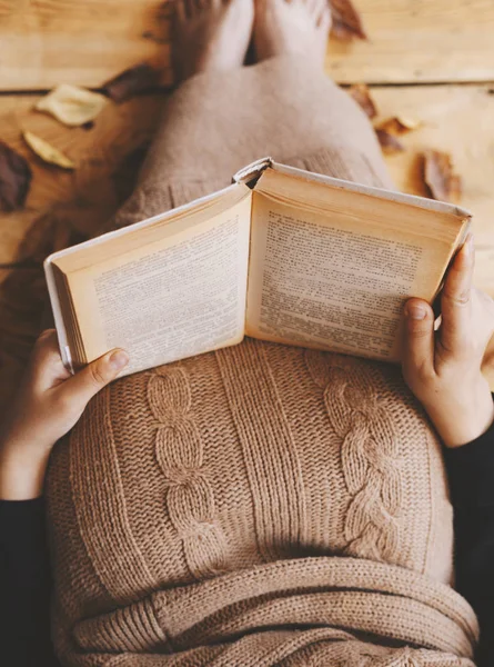 Reading book. Female sit at wooden floor with fall leaves. Woman covered with knitted plaid and reading interesting book