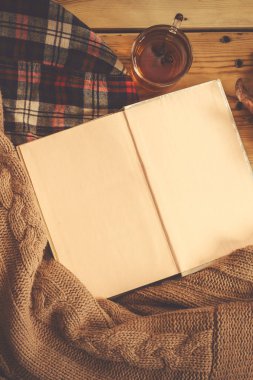 Sonbahar arka plan. Açılan kitap, sonbahar yaprağı ve örme kazak. Bir fincan sıcak çay, tahta masanın üzerine örülmüş atkı. Sonbahar ısınma içecek