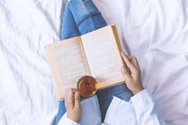 Woman on the bed with old book and cup of tea. Woman holding a book and tea in hands. Female reading book on bed in early weekend morning clipart