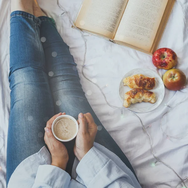 Winter holidays. Autumn morning. Woman having breakfast in bed. Coziness weekend morning. Woman with cup of coffee latte in hands, apples and croissants on bed. Relaxing Sunday activities