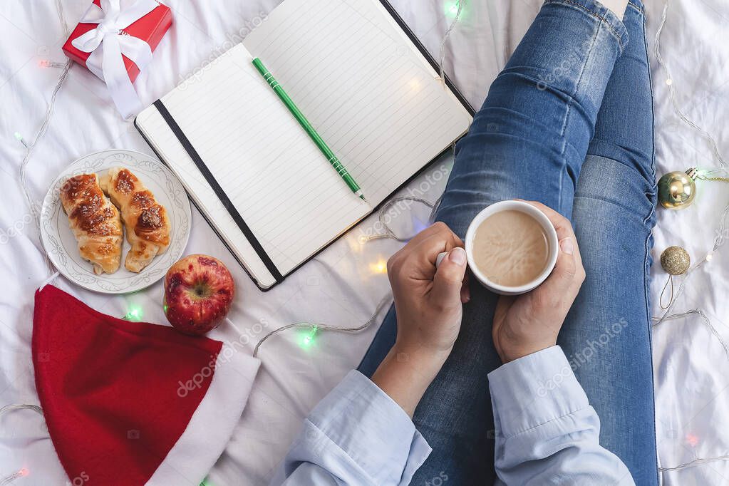 Young woman lying and having breakfast in bed. Christmas morning in a cozy bedroom decorated with Xmas lights and gifts. Writing wishes, plans and resolutions for the new year