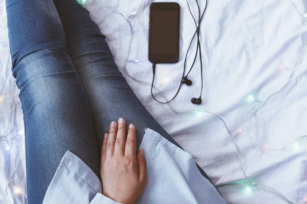 A woman trying to sleep after a hard-working day. Female relaxing in a cozy bedroom. Listening to music on a smartphone. Autumn or winter season