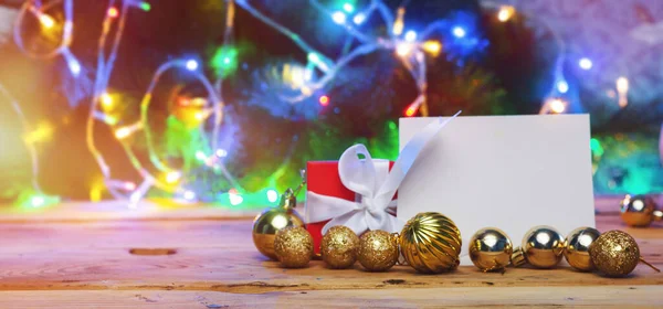 Χριστουγεννιάτικο δέντρο διακοσμήσεις και κουτί δώρου με μια λευκή κάρτα σε ένα ξύλινο τραπέζι. Χριστουγεννιάτικα παιχνίδια με λευκό χαρτί πάνω από το χριστουγεννιάτικο δέντρο και φώτα bokeh. Διακόσμηση διακοπών. Ιστορικό Xmas — Φωτογραφία Αρχείου