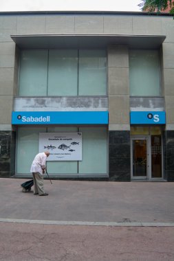 Viladecanlar, Barcelona - 2 Eylül 2025: Yaşlı bir yaya Sabadell banka şubesinin önünden geçiyor, penceresinde büyük bir reklam afişi görülüyor.