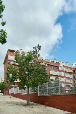 Güneşli bir günde çevresi yeşil ağaçlarla çevrili kırmızı tuğlalı ve metal çitli modern bir yerleşim yeri..