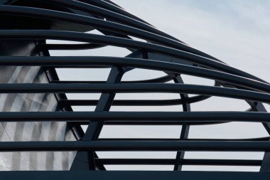 Black Metal mimari bir yapıya yakın çekim. Birbirine kenetlenmiş kavisli ve düz çubuklar soluk mavi gökyüzüne karşı dinamik bir geometrik desen oluşturur..