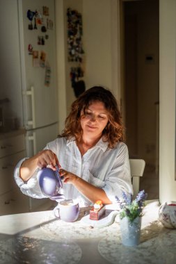 Kadın dingin bir sabah ritüeli yapar, narin bir fincana çay döker, çiçeklerle çevrili zarif bir masada, neşe ve sakinlik saçar.