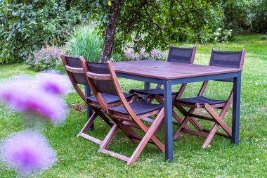 Katlanan sandalyeli ahşap yemek masası, yemyeşil ve renkli çiçeklerle çevrili canlı bir bahçede kurulmuş, rahatlatıcı ve açık havada keyif veren davetkar bir yemek masası.