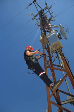 Kırmızı kasklı ve emniyet kemerli Tehnician elektrik direğinde telsiz çağrısı yapıyor.
