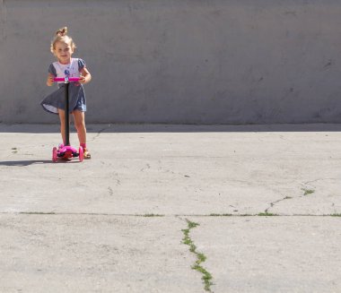 Şirin çocuk ustaca bir pembe iki tekerlekli scooter üzerinde yaz güneşli bir günde kaymaktadır.