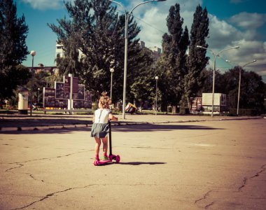 Şirin çocuk ustaca bir pembe iki tekerlekli scooter üzerinde yaz güneşli bir günde kaymaktadır.
