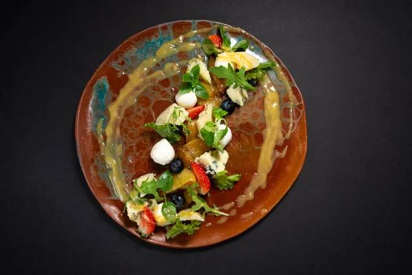 Cheese and fruit salad on a handmade plate. Mozzarella, blue cheese, parmesan, zucchini rolls, micro greens, blueberries, strawberry wedges, caramelized pear, pear puree sauce. Top view of a plate.