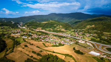 Gökyüzü manzarası yeşil yuvarlanan tepelerin arasına yerleşmiş kırsal köyü gösteriyor