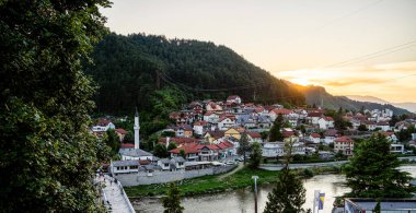 Nehir kenarındaki küçük köyü aydınlatan gün batımı yeşil ormanlık tepelerin arasına yerleşmiş.