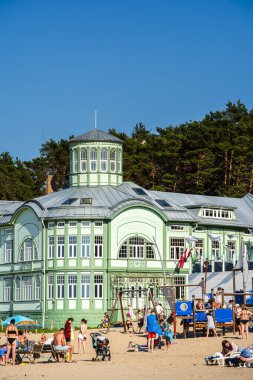 İnsanlar mavi gökyüzünün altındaki yeşil tarihi binanın yakınındaki kumsalda dinleniyorlar.