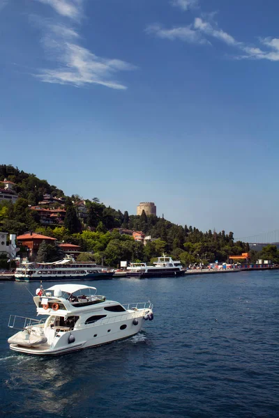 Tanınmayan insanlar İstanbul 'da Boğaziçi' nde beyaz lüks bir yata biniyorlar. Arka planda büyük motorbotlar ve Rumelihisari ya da Bogazkesen Kalesi (ortaçağ kalesi) yer alıyor. Güneşli bir yaz günü..