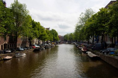 Kanal manzarası, park edilmiş tekneler, arabalar, ağaçlar ve Amsterdam 'daki geleneksel binalar. Güneşli bir yaz günü..
