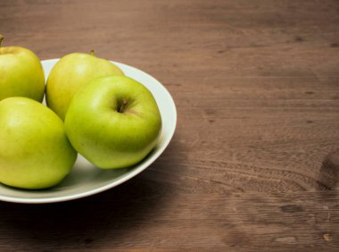 Tahta masadaki beyaz porselen kasede üç elma.