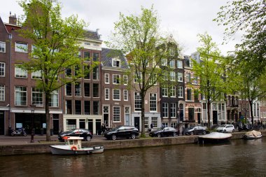 Kanal manzarası, caddelerde yürüyen insanlar, park halindeki tekneler, arabalar, ağaçlar ve tarihi yapılar, Amsterdam 'da Hollanda mimari tarzını gösteren geleneksel binalar. Bu bir yaz günü..