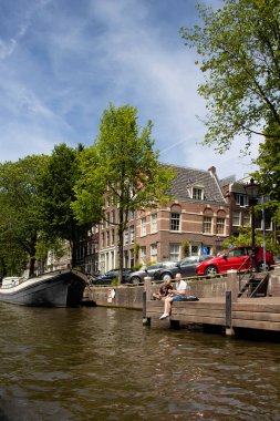 Kanalda takılan iki adamın görüntüsü, ağaçlar, park edilmiş arabalar ve bir tekne, Amsterdam 'da Hollanda mimari tarzını gösteren tarihi ve geleneksel binalar. Güneşli bir yaz günü..