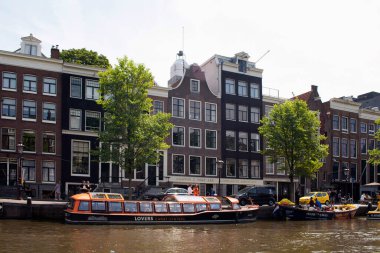 Amsterdam 'da Hollanda mimari tarzını gösteren park edilmiş kanal turu tekneleri, ağaçlar ve tarihi, geleneksel ve tipik binaların manzarası. Güneşli bir yaz günü..