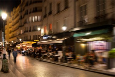 Geceleri Paris 'te kafelerin ve bistroların önünden geçen insanların bulanık görüntüsü..