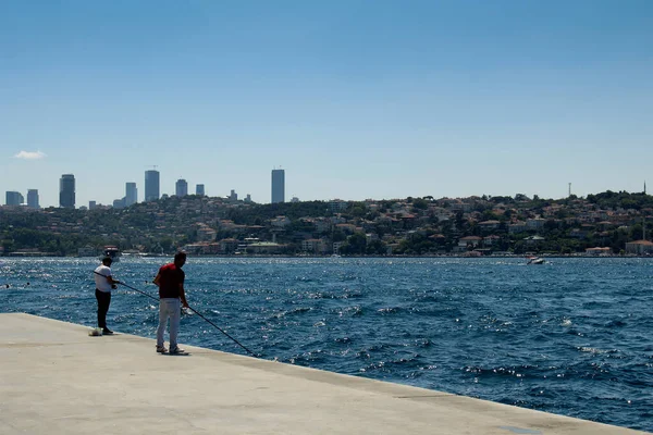 Unrecognizable two fishermen try to catch fish by Bosphorus. It is a sunny summer day in Istanbul. European side and skyscrapers are in the background.