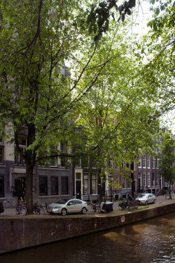 Kanal manzarası, ağaçlar, park edilmiş bisiklet ve arabalar, Amsterdam 'da Hollanda mimari tarzını gösteren tarihi ve geleneksel binalar. Güneşli bir yaz günü..
