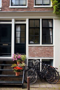 Amsterdam 'daki kültür ve yaşam tarzını gösteren tipik ve geleneksel binaların önündeki merdivenlerde park edilmiş bisiklet ve çiçek manzarası. Güneşli bir yaz günü..