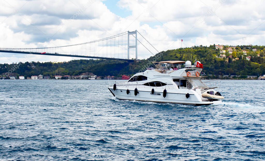 White luxury yacht / boat crosses Bosphorus between European and Asian sides in Istanbul. It is cloudy autumn day. FSM (Fatih Sultan Mehmet) bridge is in the background.