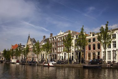 Kanal, tarihi ve geleneksel binalar, ağaçlar ve Amsterdam 'da park halindeki teknelerin orada takılan insanların görüntüsü. Mavi gökyüzüyle güneşli bir yaz günü.