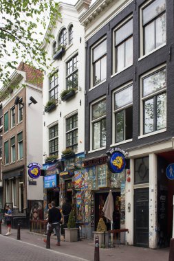 Ot kafelerden birinin manzarası ve Amsterdam 'da sokakta yürüyen insanlar. Manzarada Hollanda mimari tarzını yansıtan geleneksel ve tipik binalar da yer alıyor.