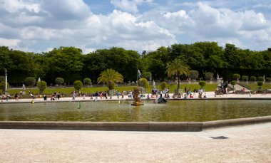İnsanlar Lüksemburg Sarayı 'nın önünde havuzun etrafında takılıyor. Havuzda küçük oyuncak yatlar ve bir çeşme var. Görüntü Paris yaşam tarzını yansıtıyor