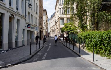 Paris 'in Le Marais semtinde bir sokakta bisiklet süren bir kadın. Binalarda yürüyen ve Fransız mimari tarzları olan insanlar görüyoruz.