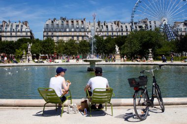 Erkek ve kadın Jardin Des Lüksemburg 'da havuz başında yemek yiyorlar. Paris 'te bir bisiklet, dönme dolap, çeşme ve parkın yanındaki binalar görülüyor.