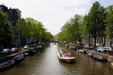 Bir gezi kanalı tur teknesi, ağaçlar, park etmiş arabalar ve botlar Amsterdam 'da Hollanda mimari tarzını gösteren tarihi ve geleneksel binalar. Güneşli bir yaz günü..