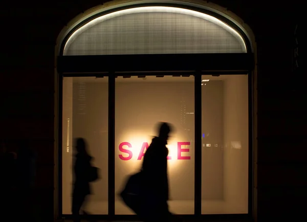 Silhouettes of man and woman walking in blurry motion in front of a fashion store's window in night. Conceptual image of sale season, consumerism and capitalism of modern world.