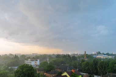 Dark storm clouds and rainfall cover a city landscape with buildings and trees, as the sky brightens slightly near the horizon.