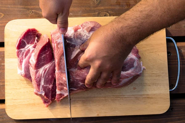 Overhead view of raw piece of pork on wooden background. Piece of fresh boneless pork, neck part or collar. Big piece of red raw meat. Butcher man hands close up: cutting meet in wooden board